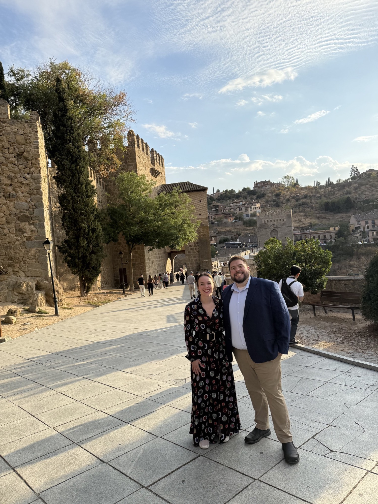 Two travelers on a stone walkway beside the medieval walls of Toledo, Spain.